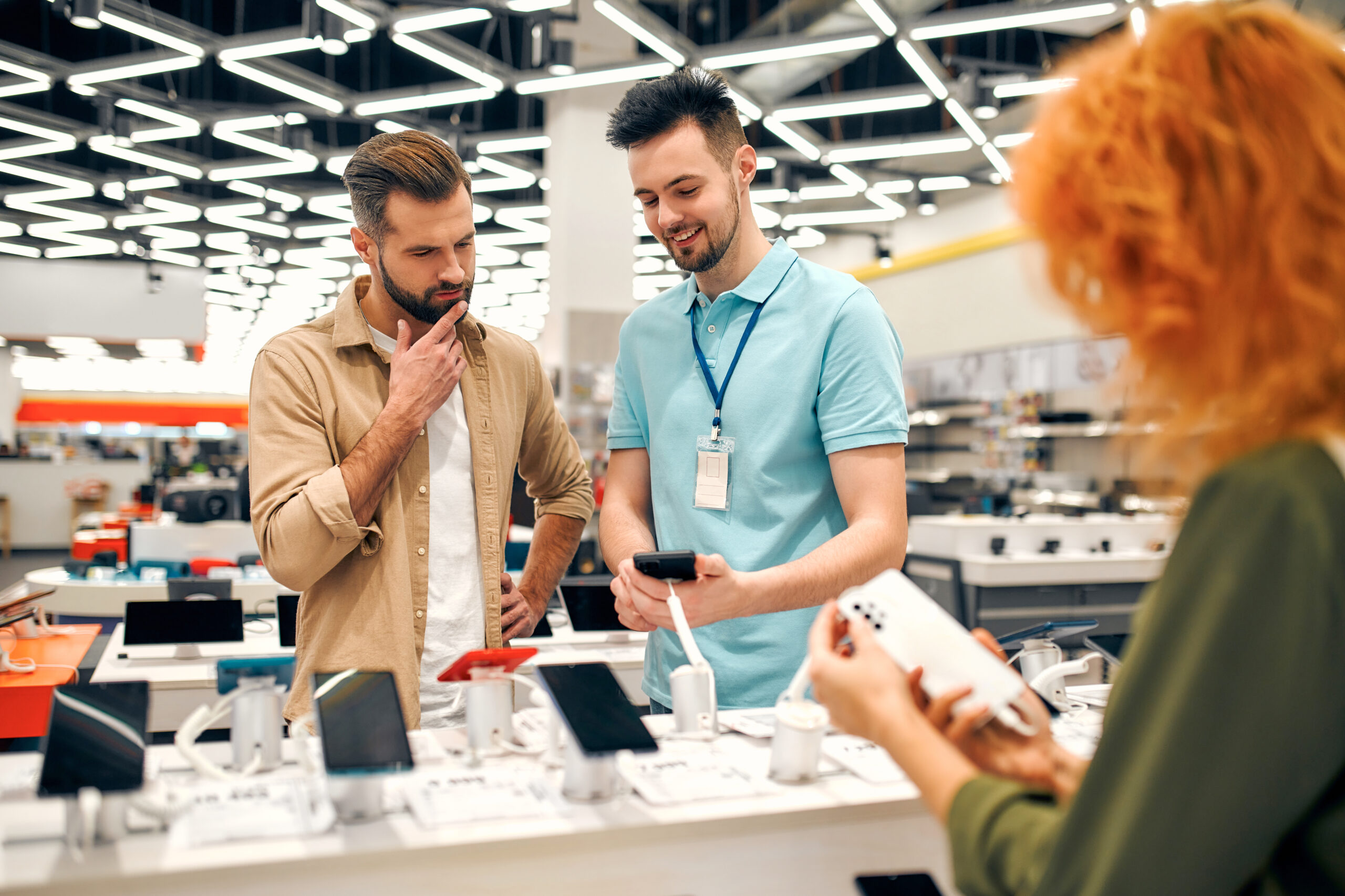 A man in a beige shirt is thoughtfully looking at a smartphone in his hand, guided by a store employee wearing a light blue polo shirt and a lanyard. The setting appears to be an electronics store with various smartphones displayed on a counter. Another person with orange hair, visible from behind, is also holding a smartphone, examining it. The store is brightly lit with modern, geometric lighting on the ceiling.