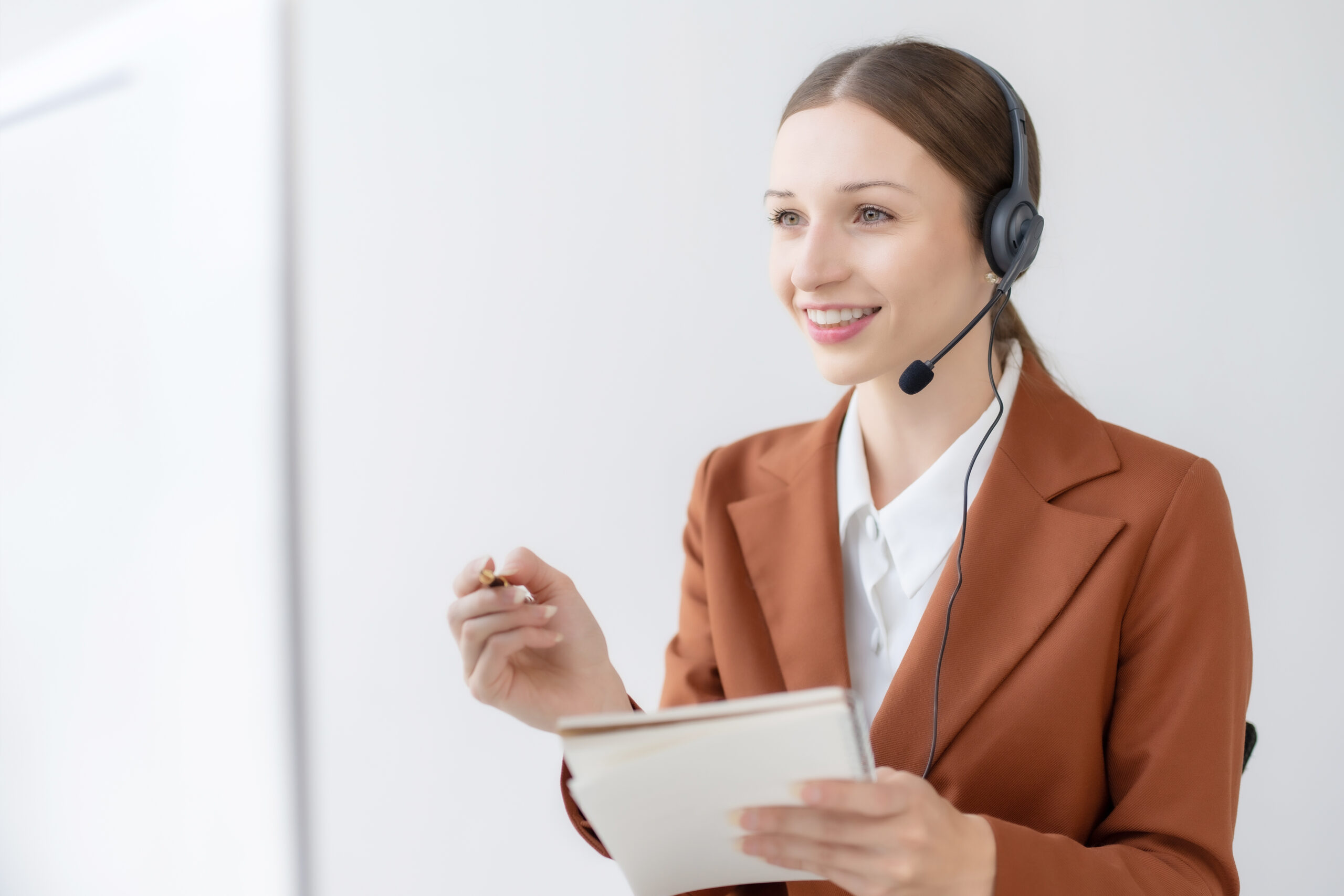 A customer service representative wearing a brown blazer and a white shirt is seated, holding a notepad and pen in her hands. She is smiling and wearing a headset with a microphone, suggesting she is engaged in a conversation or providing customer support. The background is simple and white, keeping the focus on her.