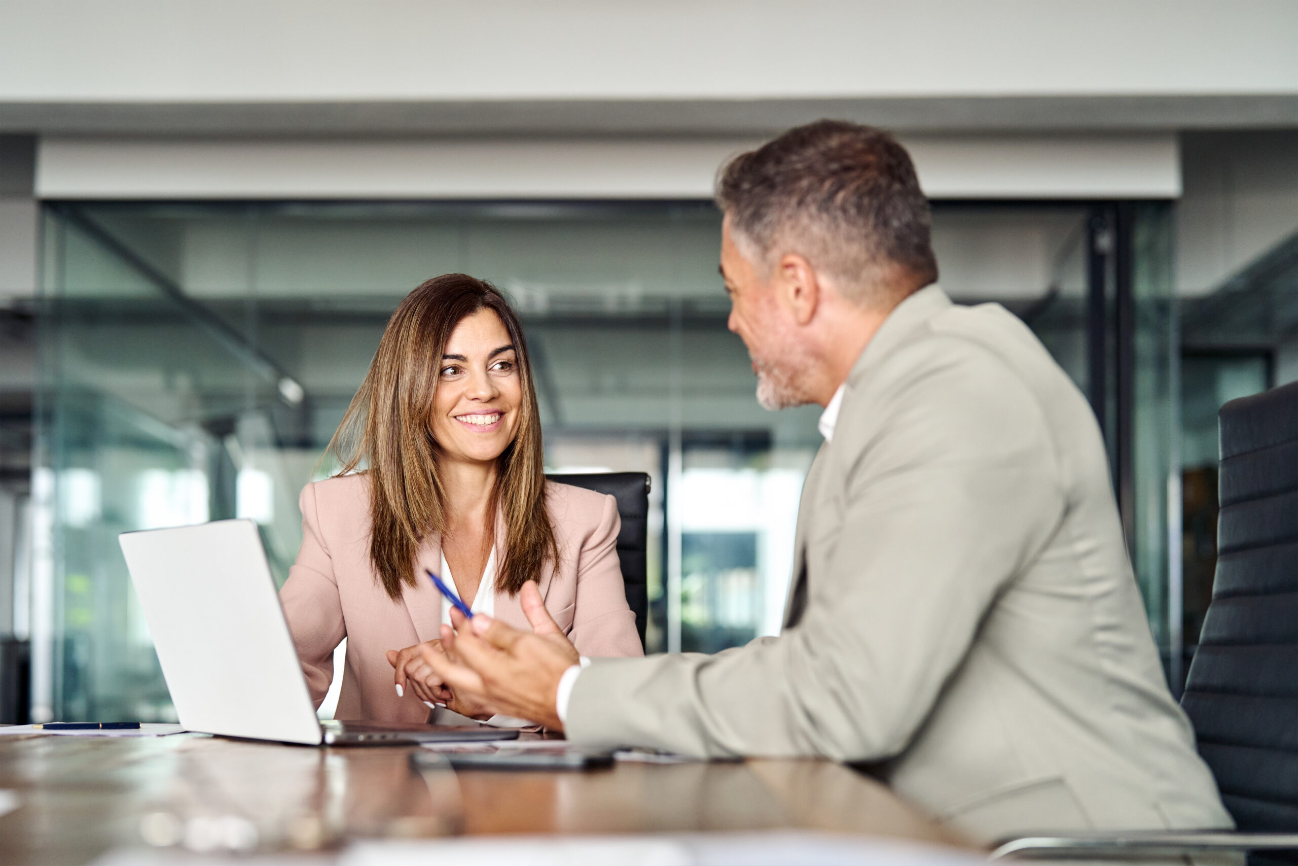 The image shows two professionals, a woman and a man, engaged in a conversation in a modern office setting. The woman, wearing a light pink blazer, is smiling and looking at the man, who is dressed in a light-colored suit. They appear to be discussing something, with a laptop and some papers on the table in front of them. The background includes glass walls and a bright, open office environment.