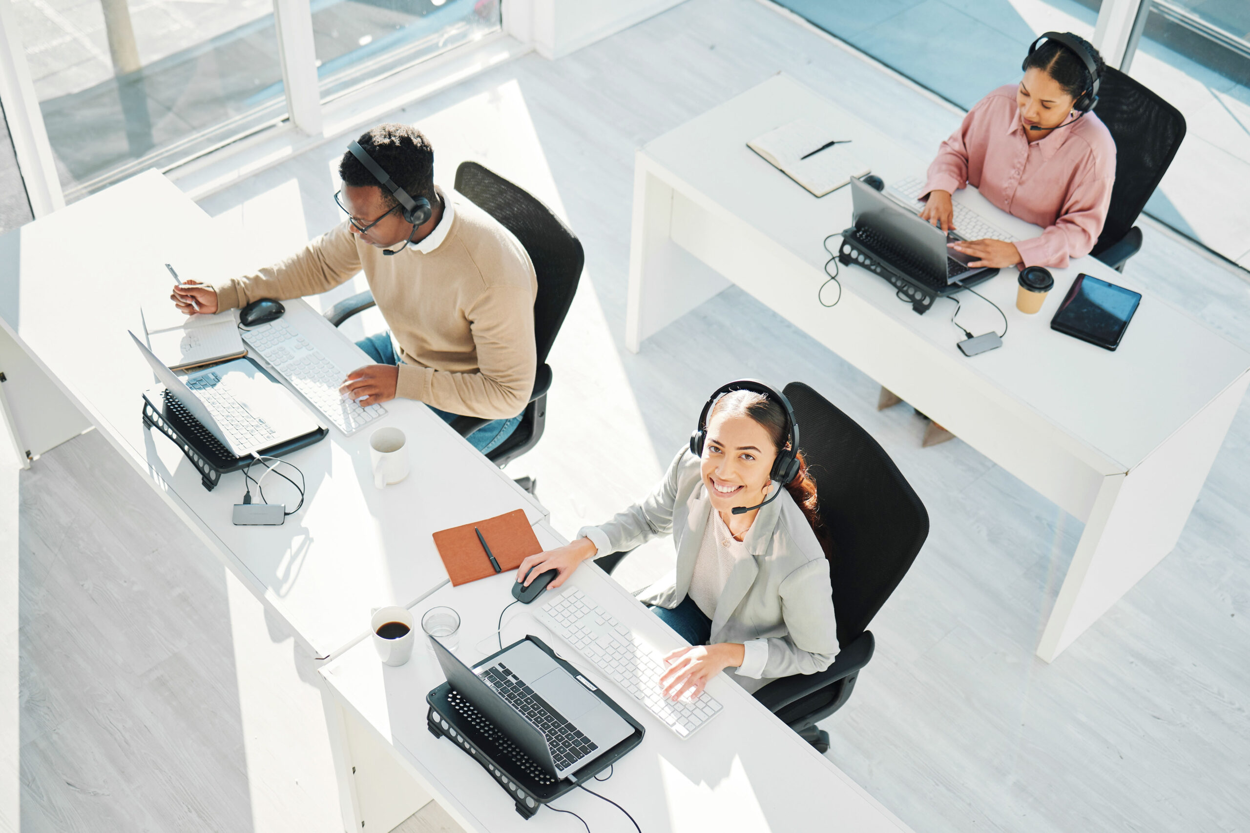 Three contact center agents working at their desks, each equipped with a laptop and headset. One agent, in the foreground, is smiling and looking up at the camera. The work environment is bright and modern, with large windows letting in natural light