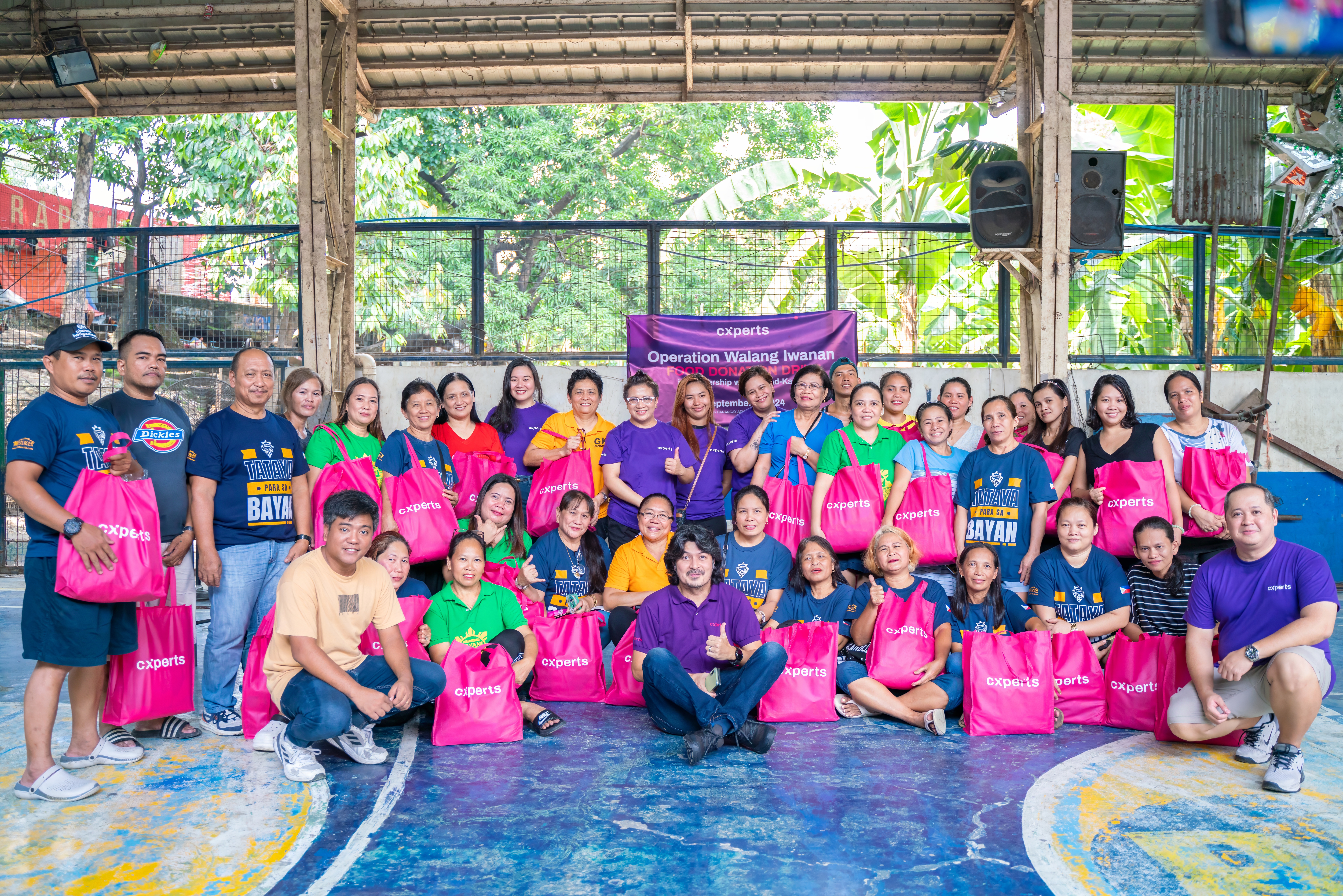 A group photo of Cxperts employees and community members during the 'Operation Walang Iwanan' CSR event. The team is seen holding pink donation bags, smiling in a sports court, underlining Cxperts' commitment to community support and customer experience. A banner in the background highlights the event's theme of partnership and giving back