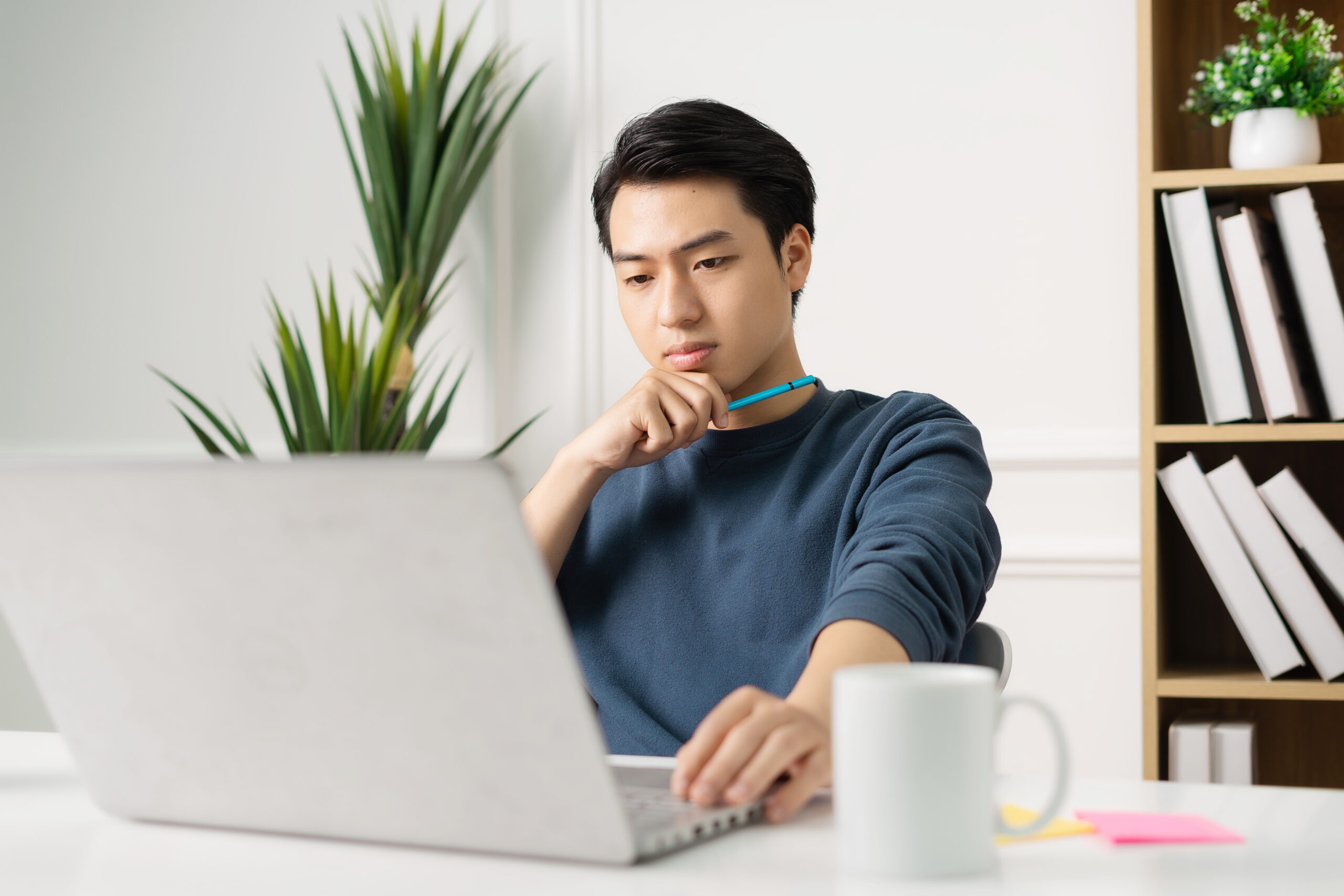 The image shows a young man sitting at a desk, focused on working on his laptop. He is holding a pen near his chin, appearing deep in thought as he looks at the screen. The setting appears to be a modern, well-lit office or study area, with a small plant to the side and shelves filled with books in the background. A white mug and a few sticky notes are placed on the desk beside him.