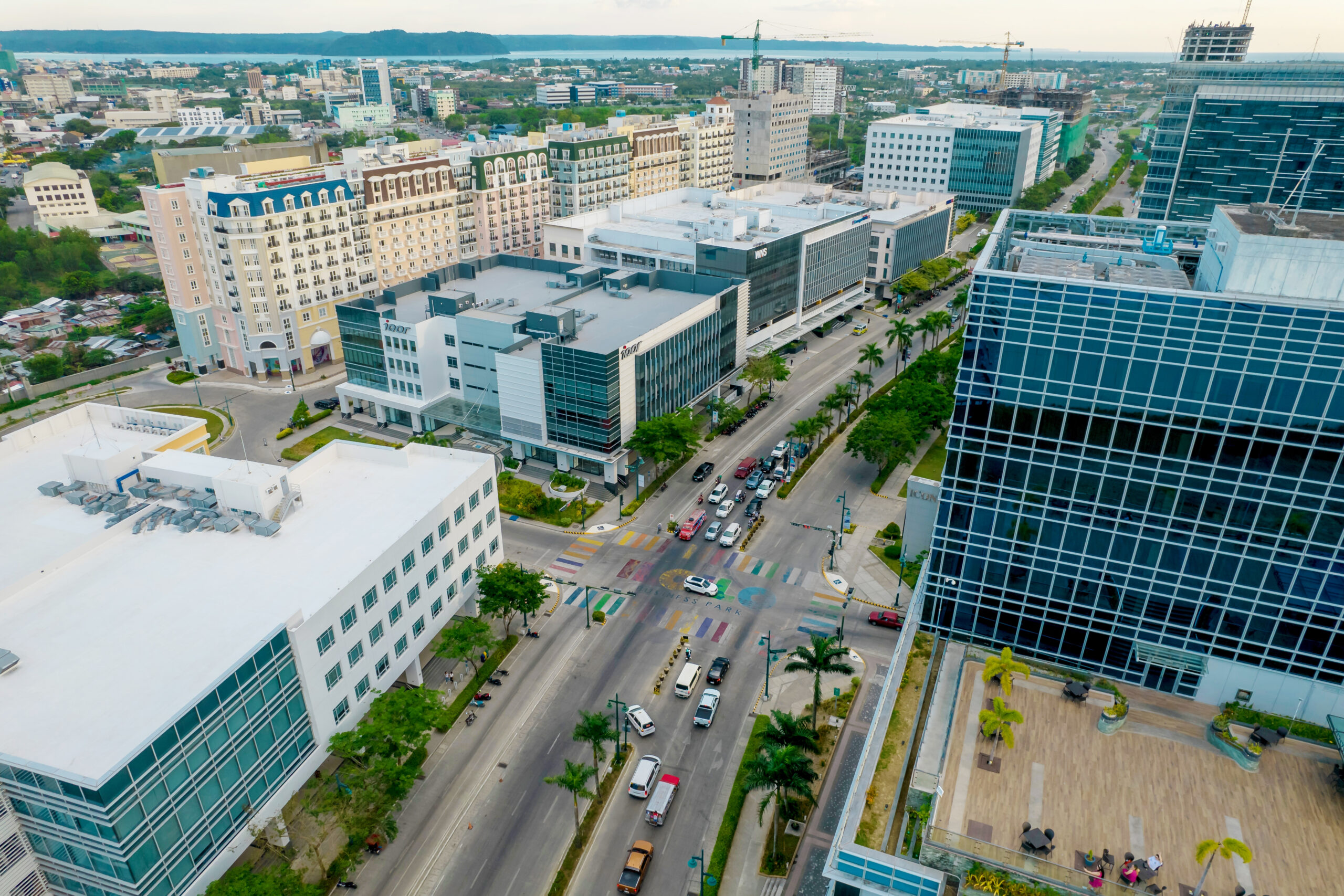 Aerial view of a modern urban area with multi-story buildings, palm-lined streets, and colorful pedestrian crosswalks.