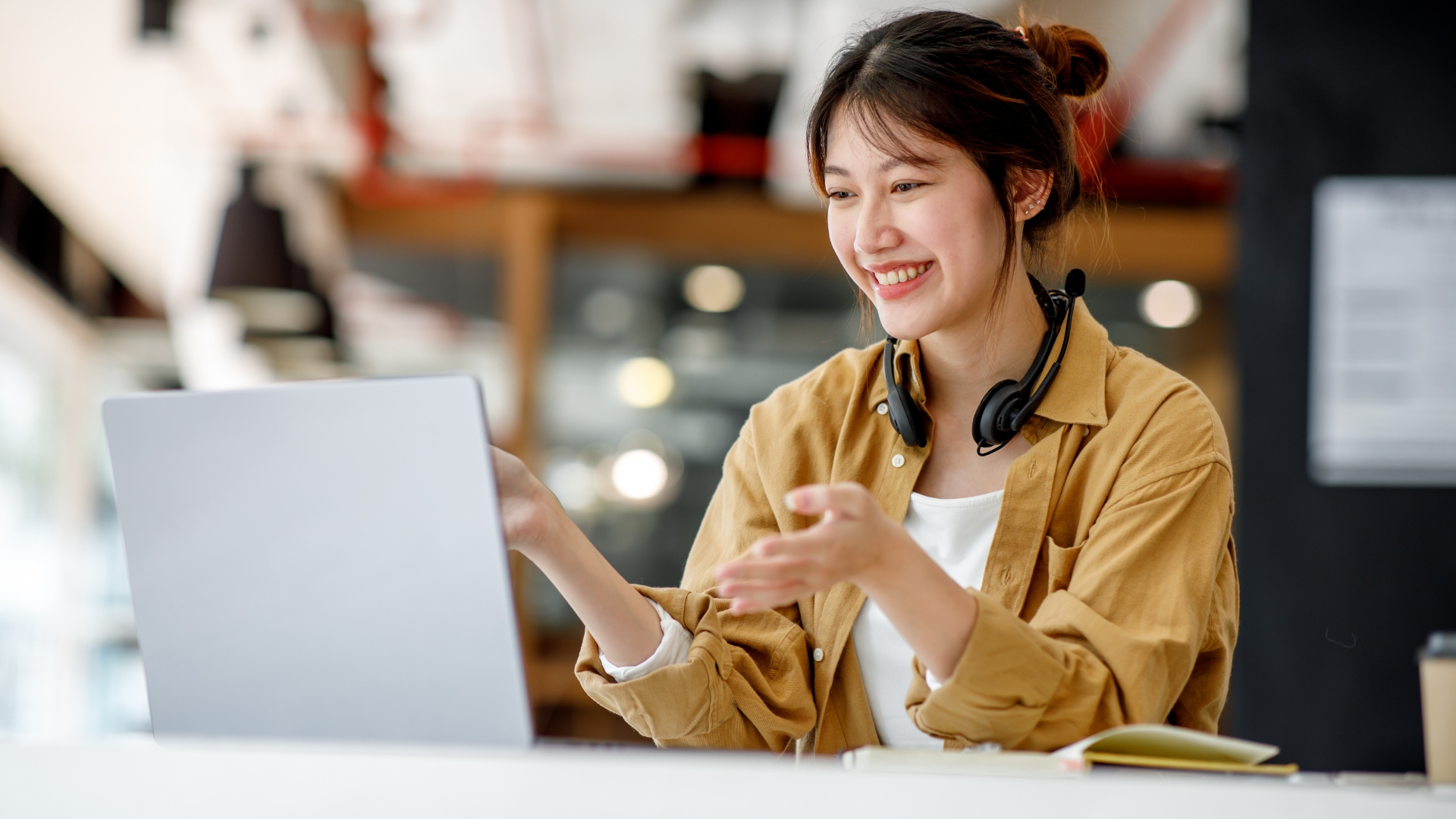 A smiling woman wearing headphones, seated at a desk in a modern workspace, gesturing while interacting with a laptop. The scene reflects a professional environment, showcasing engagement and innovation, aligning with Cxperts' focus on AI-driven customer experience solutions