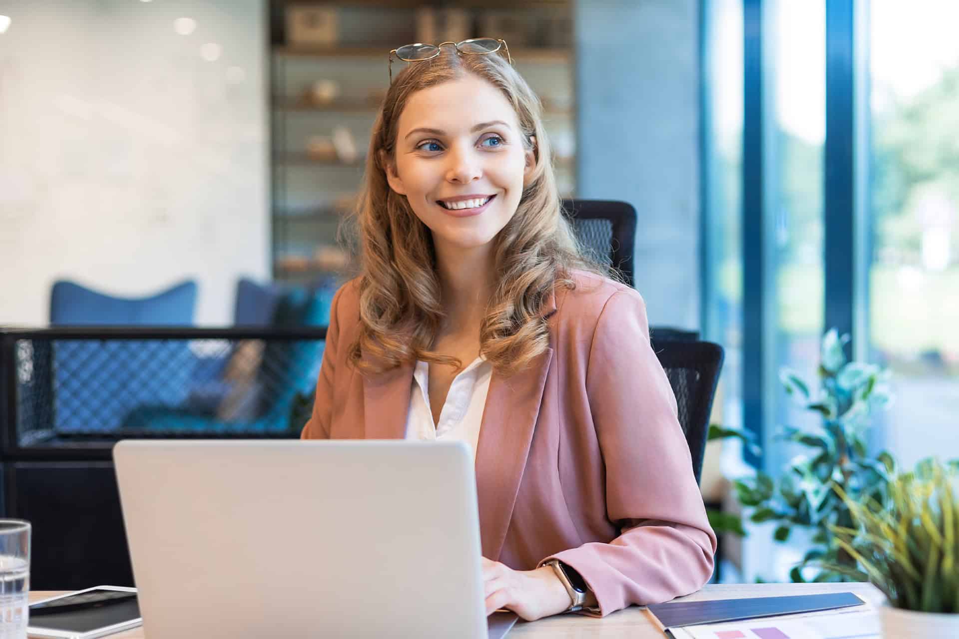 A woman in a pink blazer sits at a desk with a laptop, smiling and looking to the side. The setting is a bright, modern office with greenery.