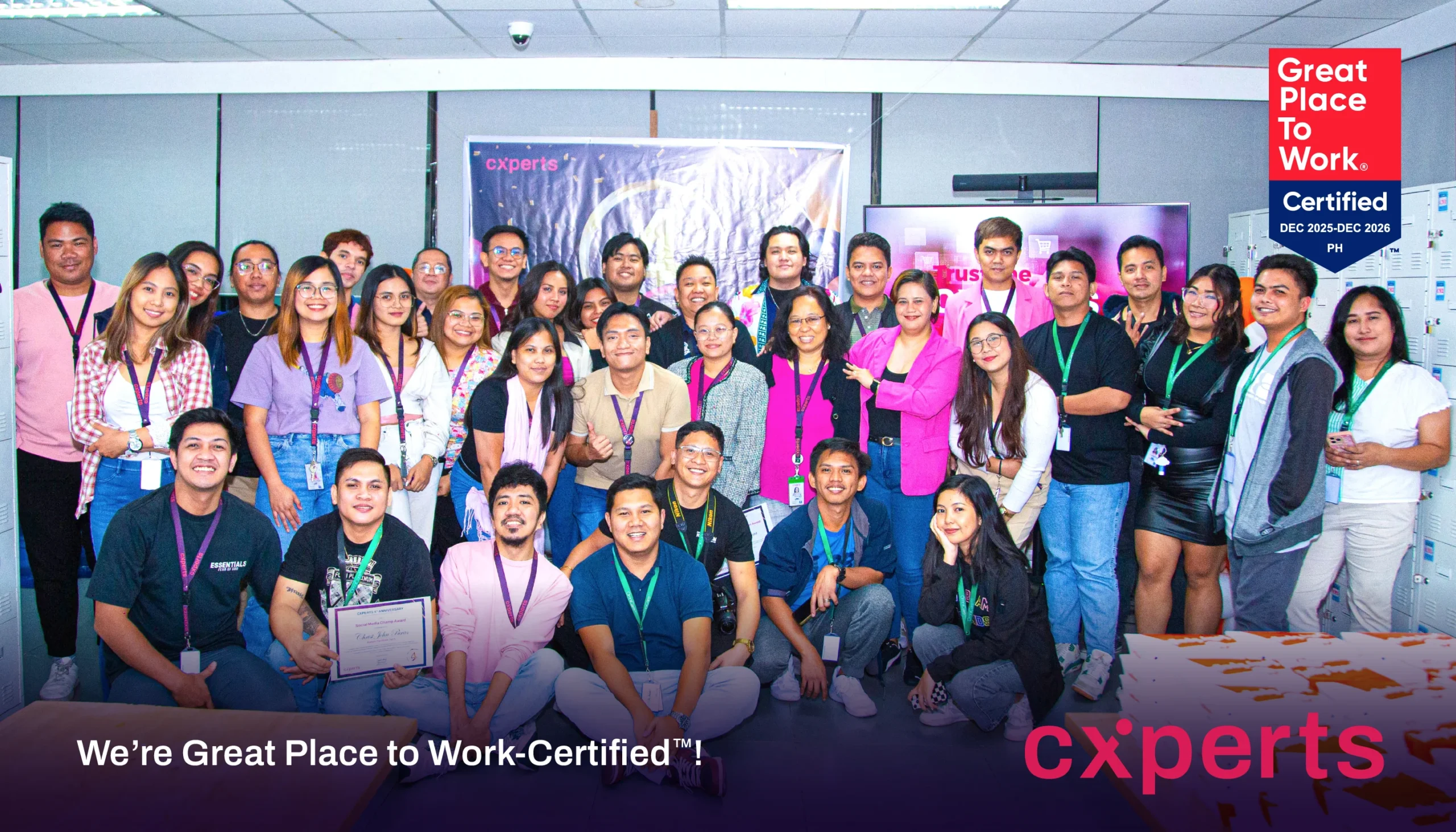 A large, diverse group of smiling employees poses together in an office setting. A "Great Place to Work-Certified" sign is visible, conveying pride and accomplishment.