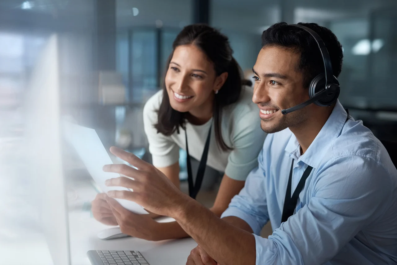 Man with headset and woman smiling, collaborating at a computer in an office. The scene conveys teamwork, technology, and a positive work environment.