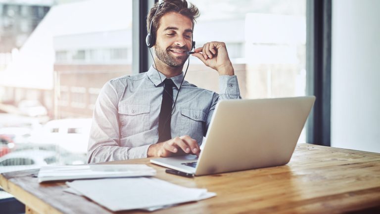 A professional man wearing a headset and working on a laptop, smiling while interacting with a customer. The scene is set in a modern office with large windows, suggesting a focus on customer experience (CX) and remote communication technology.