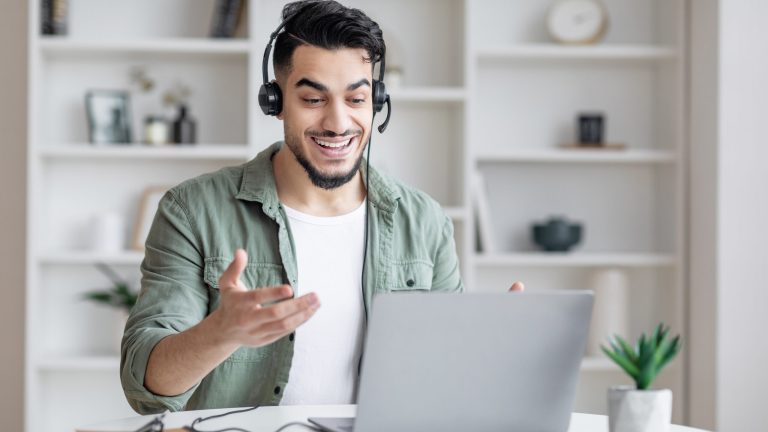 A smiling man with a beard, wearing a green shirt and headset, is gesturing during a video call on his laptop. White shelves with books and decorations are in the background.