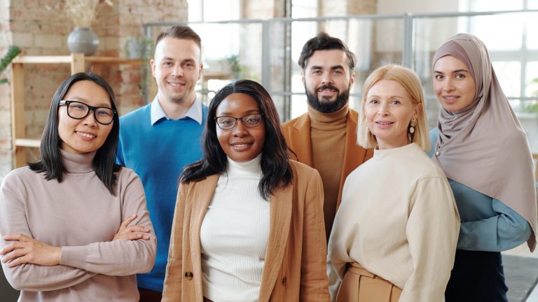 A diverse group of six professionals in business casual attire standing together in a modern office with exposed brick walls and large windows