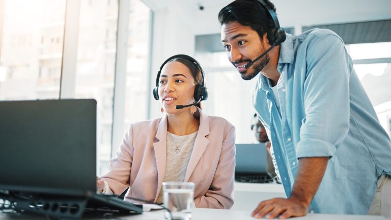 Two professional agents wearing headsets are looking at a laptop and discussing something in a bright, modern office.