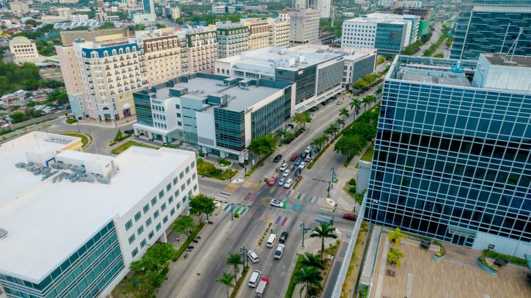 Aerial view of a modern urban area with multi-story buildings, palm-lined streets, and colorful pedestrian crosswalks.