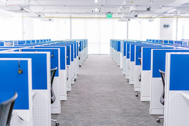 A brightly lit office space with multiple cubicles arranged in rows. Each cubicle is equipped with a computer, keyboard, phone, and headset, surrounded by blue partition walls. The area has a clean, modern look with gray carpet flooring and ergonomic chairs