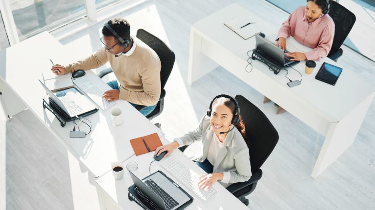 Three contact center agents working at their desks, each equipped with a laptop and headset. One agent, in the foreground, is smiling and looking up at the camera. The work environment is bright and modern, with large windows letting in natural light