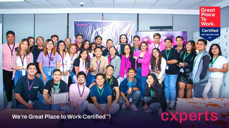 A large, diverse group of smiling employees poses together in an office setting. A "Great Place to Work-Certified" sign is visible, conveying pride and accomplishment.