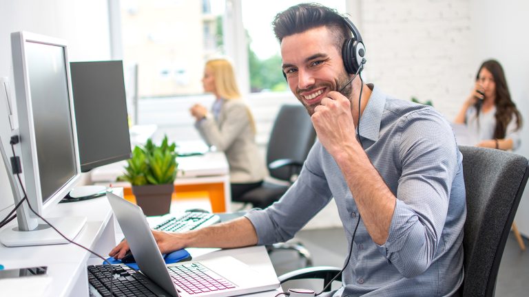 Smiling call center agent wearing a headset working on a laptop in a modern BPO office, representing positive workplace culture and employee engagement.