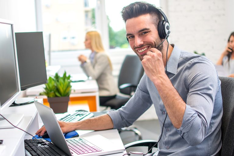 Smiling call center agent wearing a headset working on a laptop in a modern BPO office, representing positive workplace culture and employee engagement.