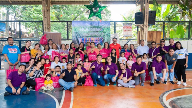 Group photo of cxperts volunteers with Gawad Kalinga Housing Project beneficiaries during a community outreach event, showing smiles and distribution of pink cxperts gift bags