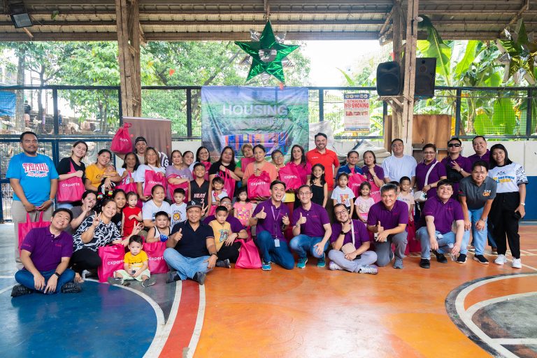 Group photo of cxperts volunteers with Gawad Kalinga Housing Project beneficiaries during a community outreach event, showing smiles and distribution of pink cxperts gift bags